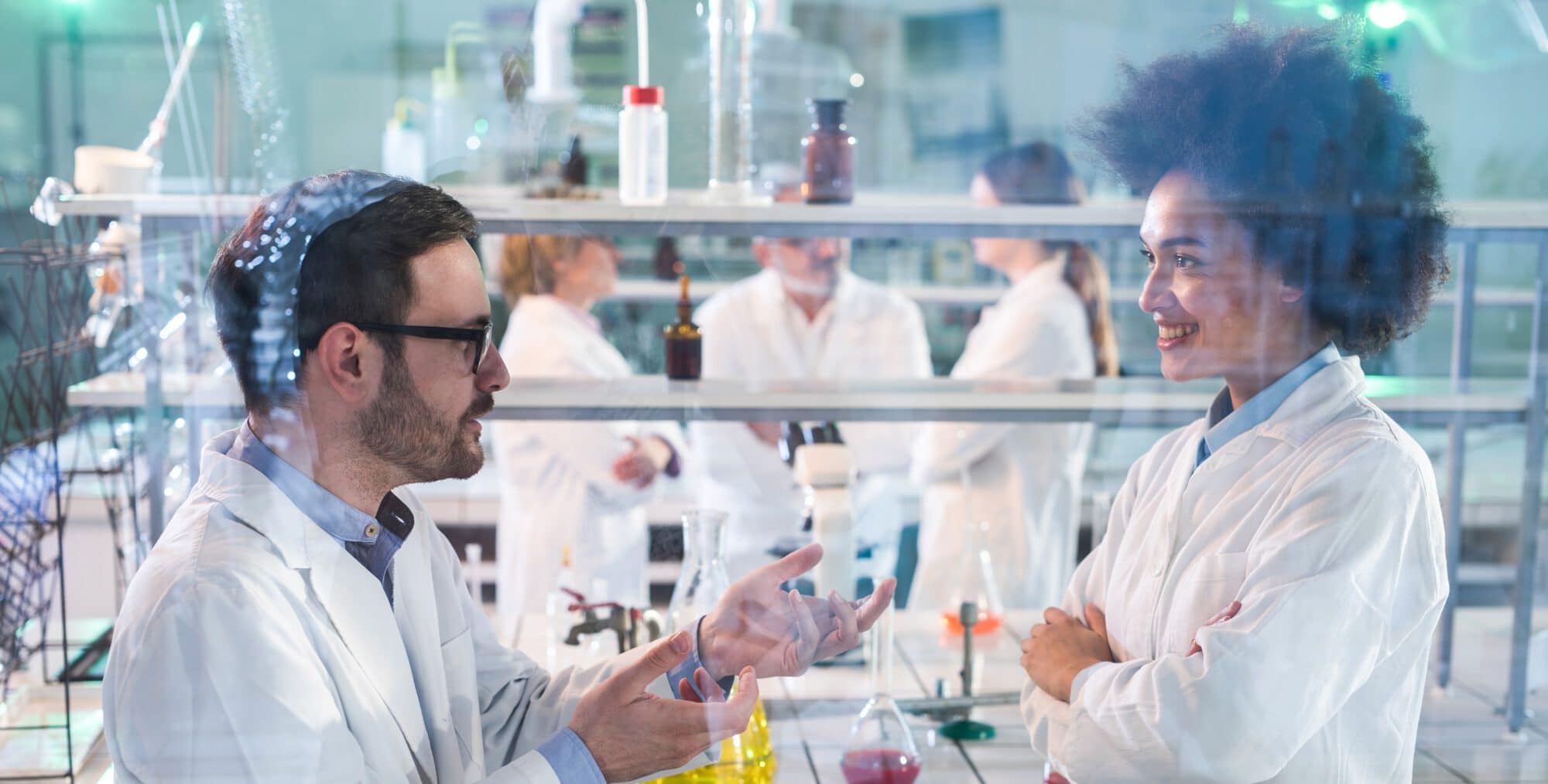 Two scientists discussing about research while working in laboratory. There are people in the background. The view is through glass.